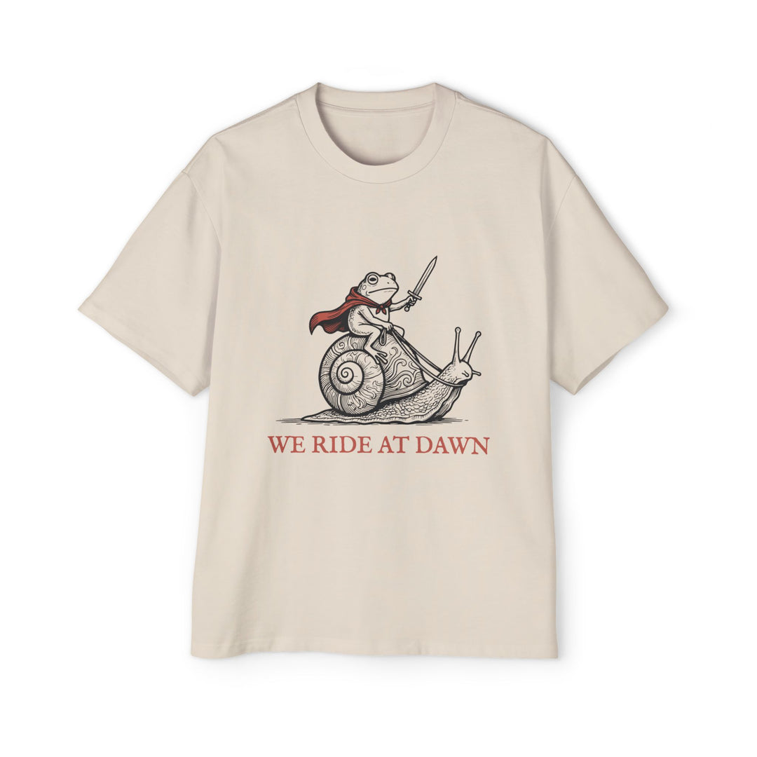 Men's natural cream graphic t-shirt featuring a whimsical illustration of a frog wearing a flowing red cape and riding a large snail while holding a sword. The vintage-inspired black line art design includes the phrase "WE RIDE AT DAWN" beneath the artwork, creating a humorous fantasy and cottagecore aesthetic.